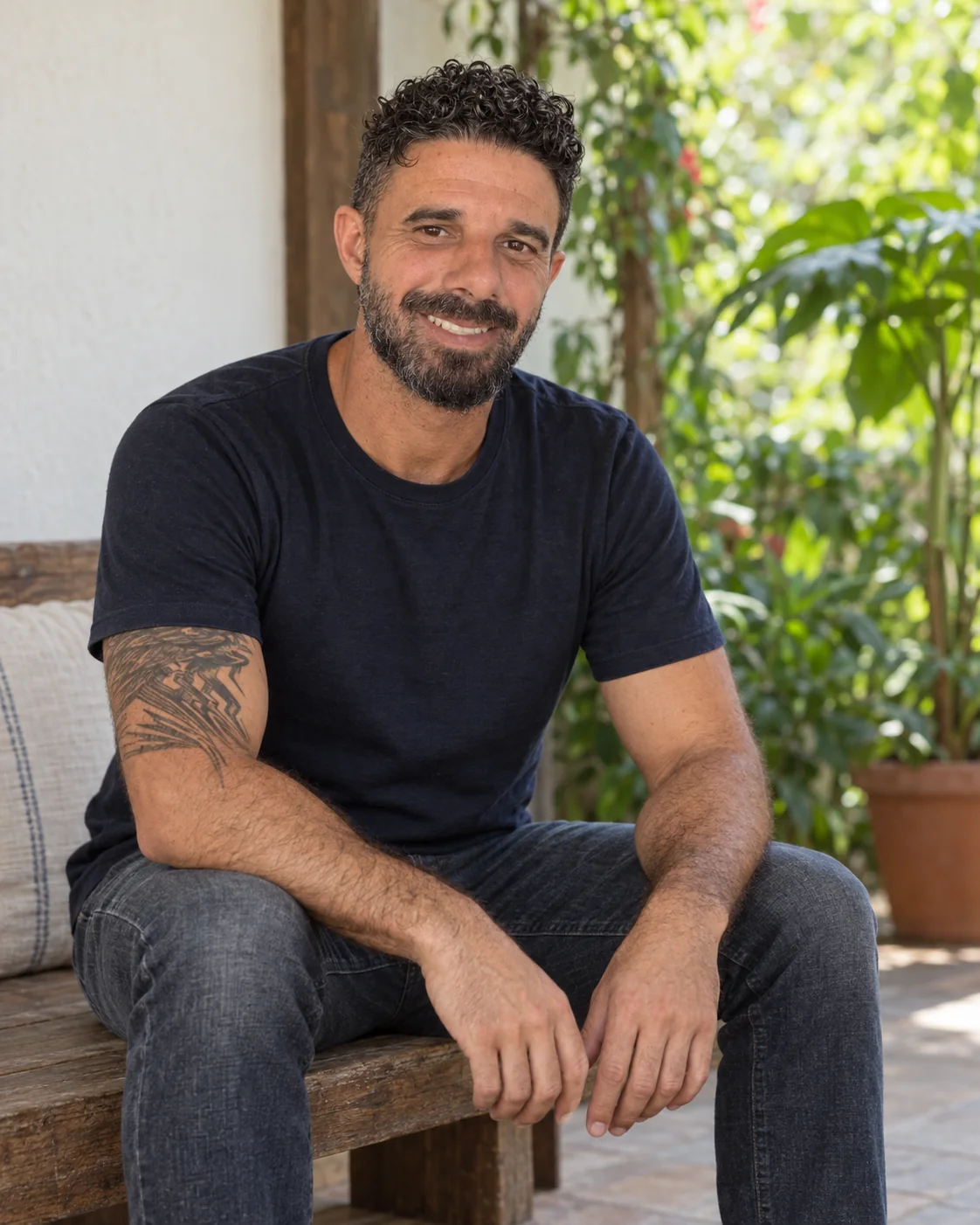 Bruno de camiseta azul-marinho e jeans, sentado em um banco junto ao jardim, sorrindo para a câmera